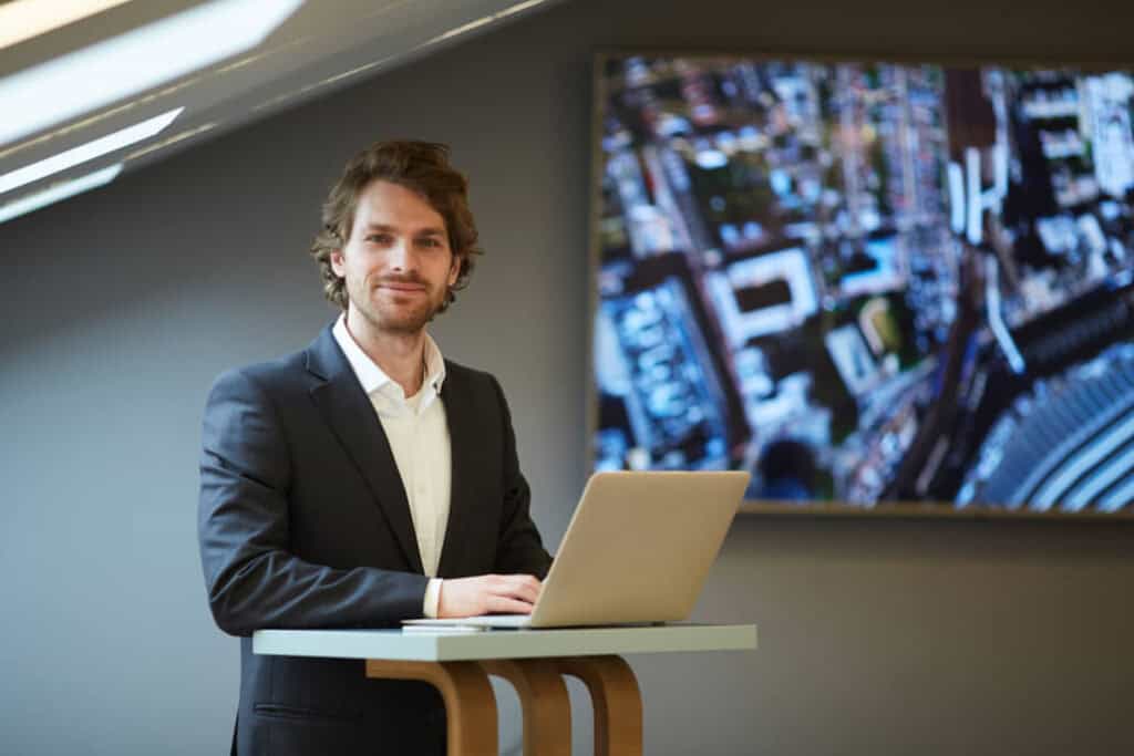 A man in a suit is standing at a high table with an open laptop. He is in a modern office setting, and a blurred, abstract image is displayed on a large screen in the background.