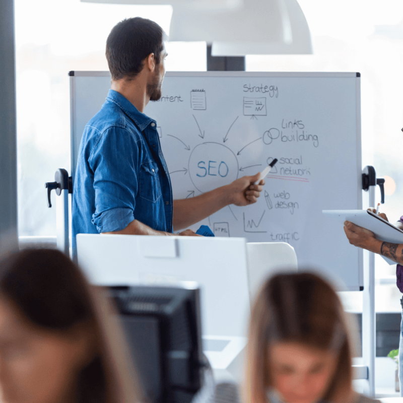 Two people stand by a whiteboard discussing SEO for dentists. One points at the board with a marker while the other writes on a clipboard. Other people are blurred in the foreground, highlighting an office setting.